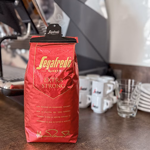eine Packung Segafredo Extra Strong verpackt mit Aromaklammer vor einer Kaffeemaschine und einigen Tassen